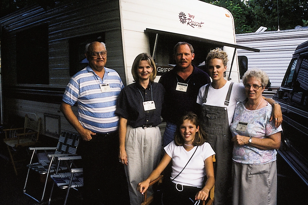 Six adults, one seated, pose beside a recreational vehicle and outdoor camping equipment.