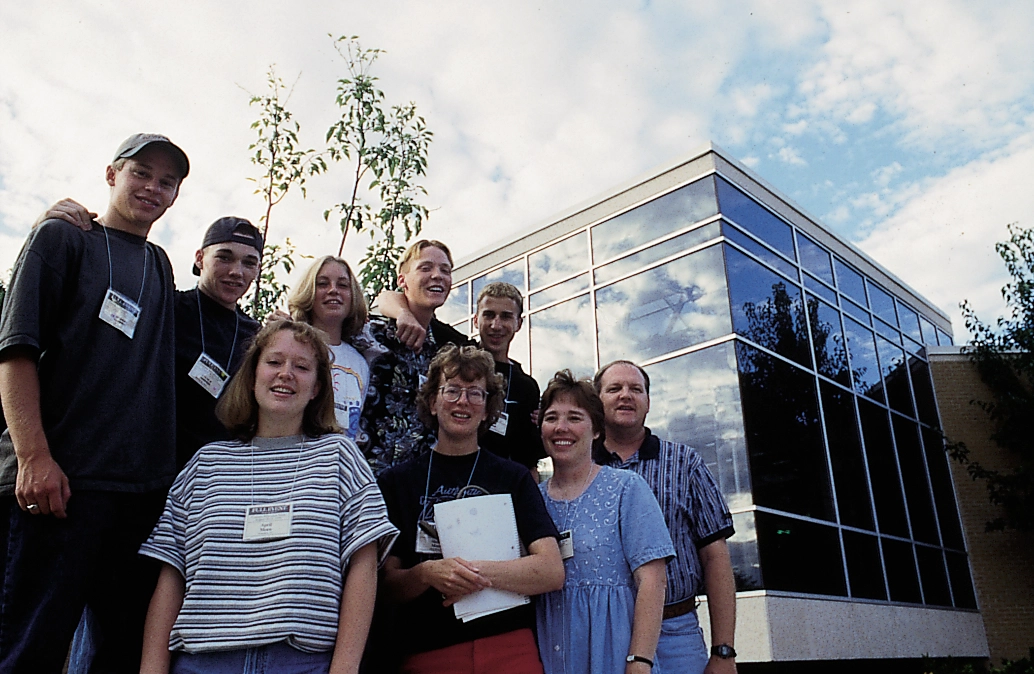 A group of adults wearing conference name tags poses outside a modern glass-fronted building.