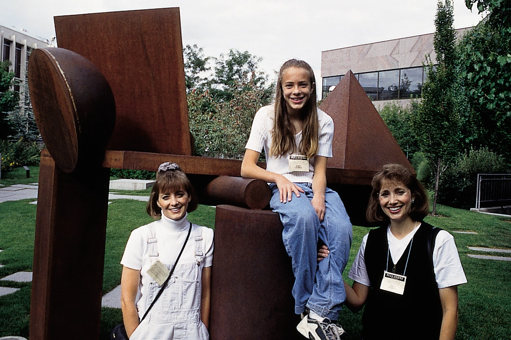 Three women wearing name tags pose beside and atop a large rust-colored abstract metal sculpture.