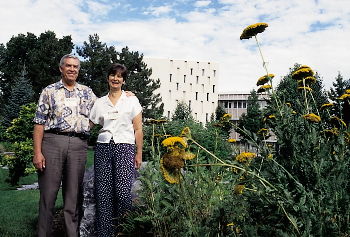 An older couple wearing name tags stands among tall flowering plants, with a campus building in the distance.