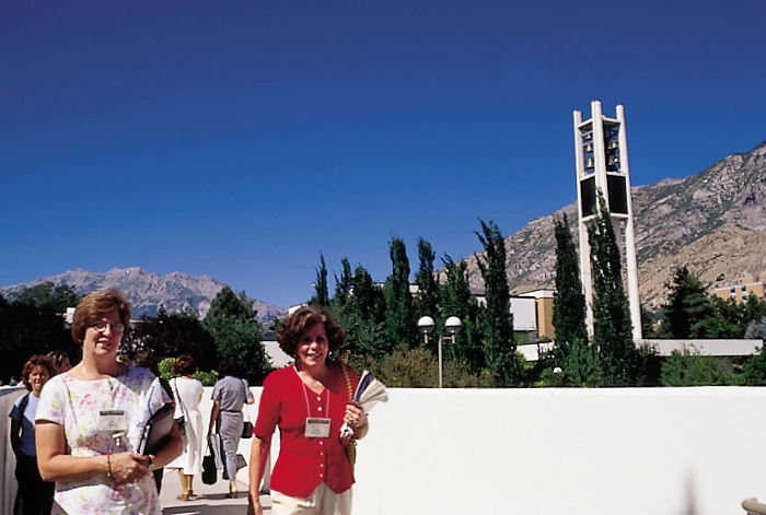 Two women wearing conference name tags stand outdoors with a campus bell tower and mountains behind them.