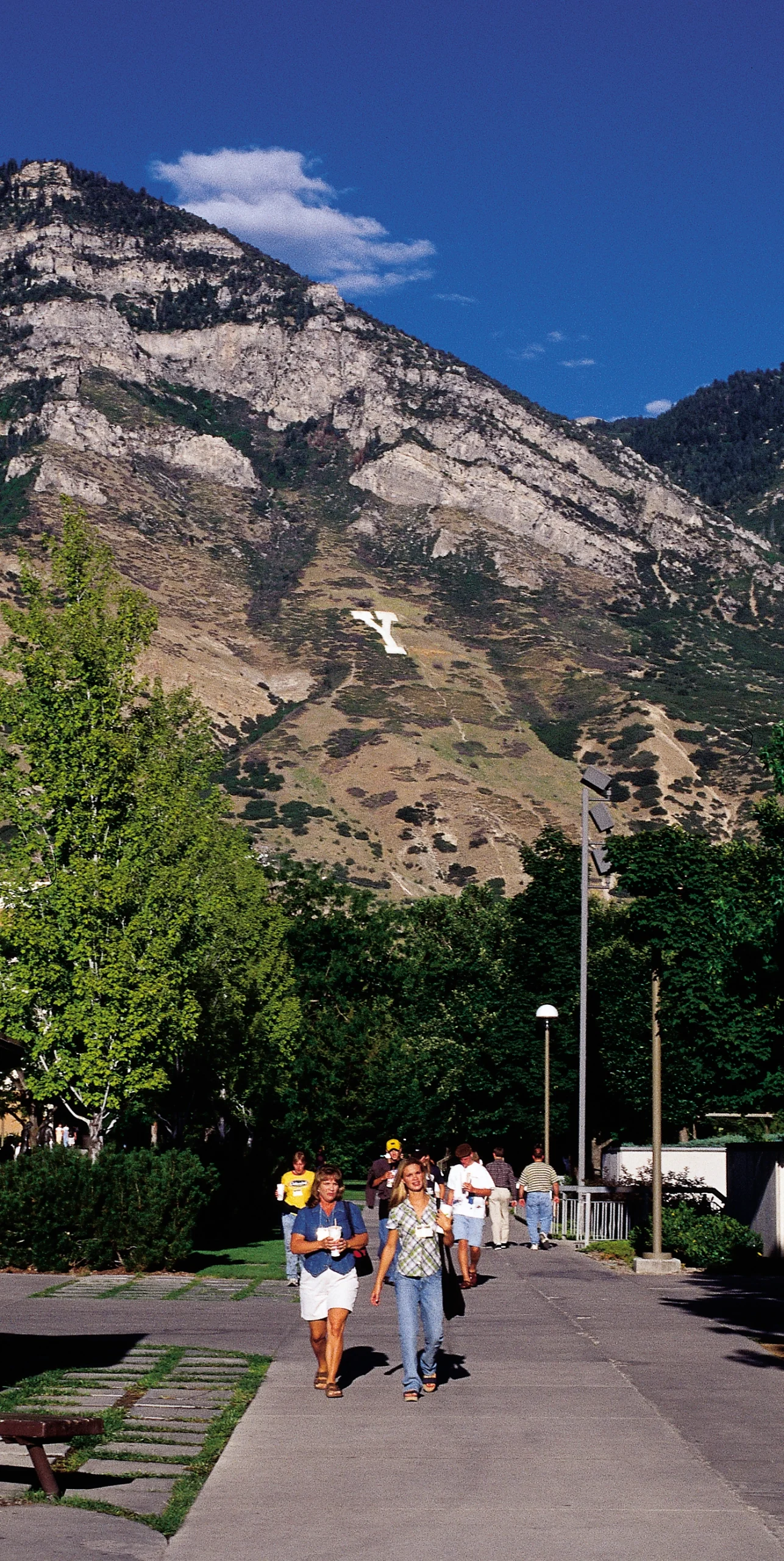 Education Week attendees walk along a tree-lined campus path beneath a steep mountain and clear blue sky.