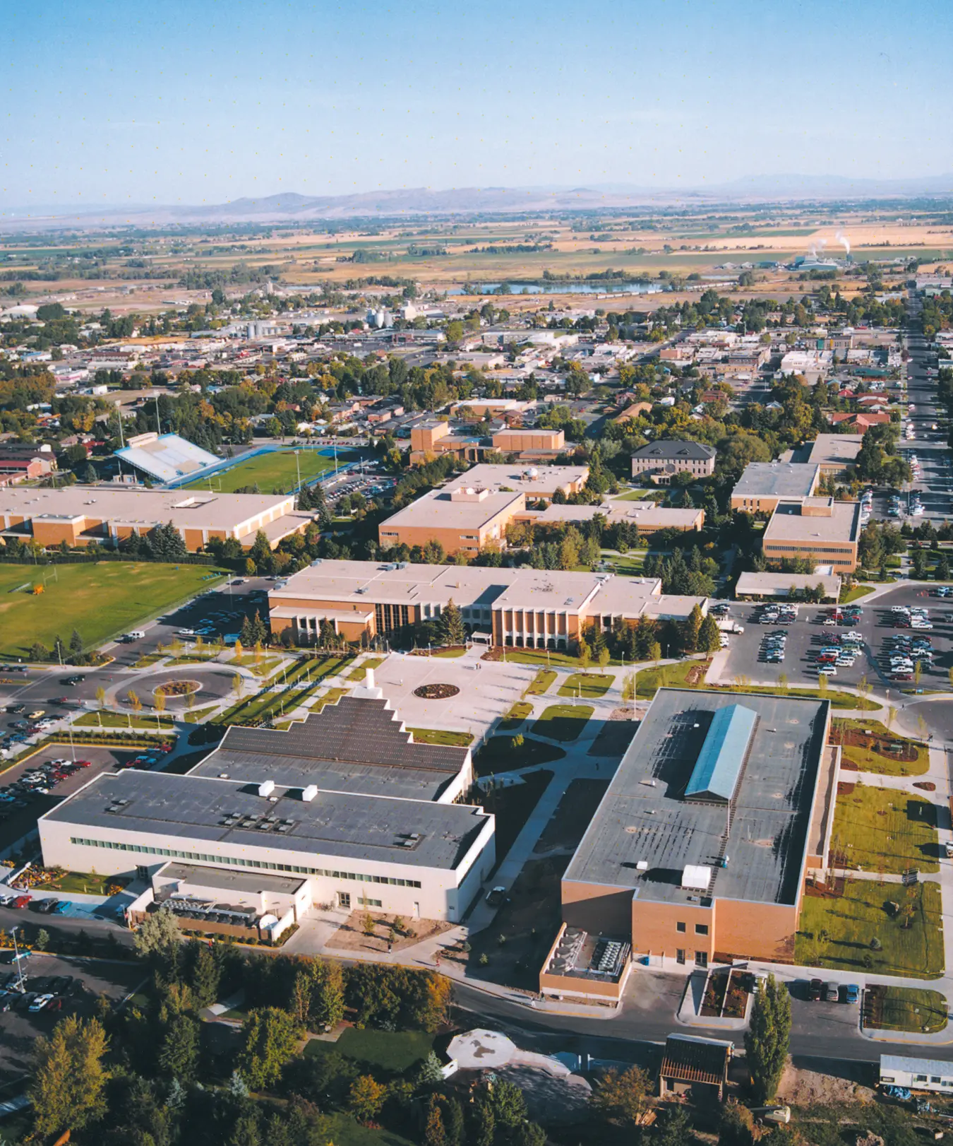 Color aerial view of the expanded BYU–Idaho campus and surrounding Rexburg, with academic buildings, athletic fields, landscaped grounds, neighborhoods, and farmland stretching into the distance.