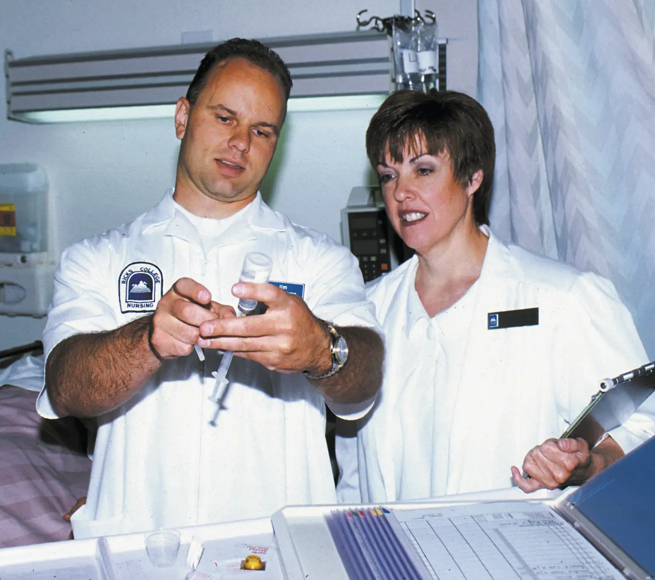 Two people in white uniforms work together in a laboratory or clinical classroom; one holds a small piece of equipment while the other watches.