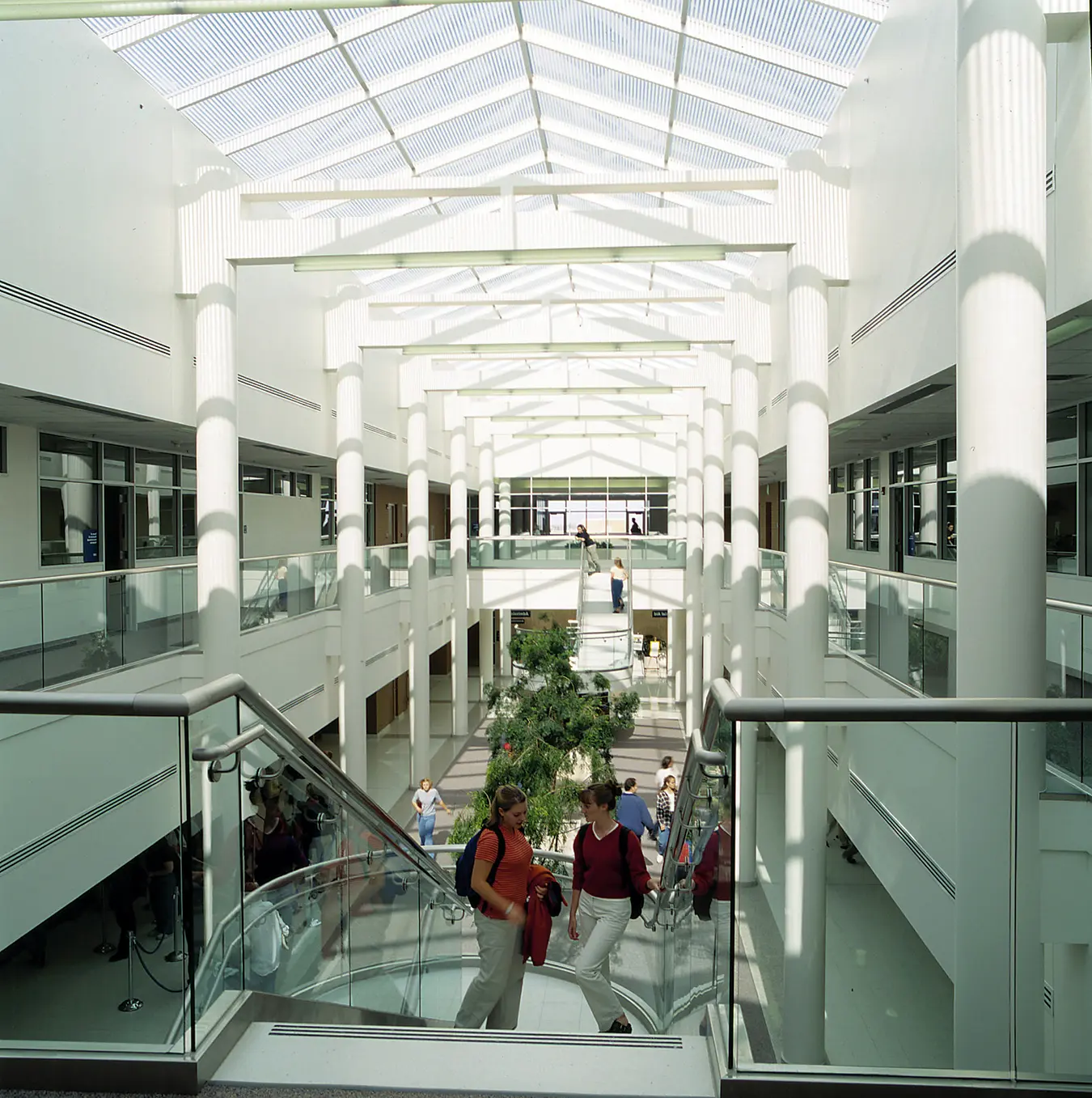 Students walk up a broad staircase inside a bright, multi-level BYU–Idaho building with white columns, skylights, glass railings, and an open central atrium.