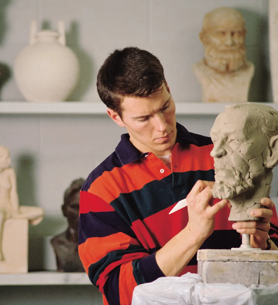 Student in a red-and-navy striped shirt carefully works on a sculpted human head in an art studio.