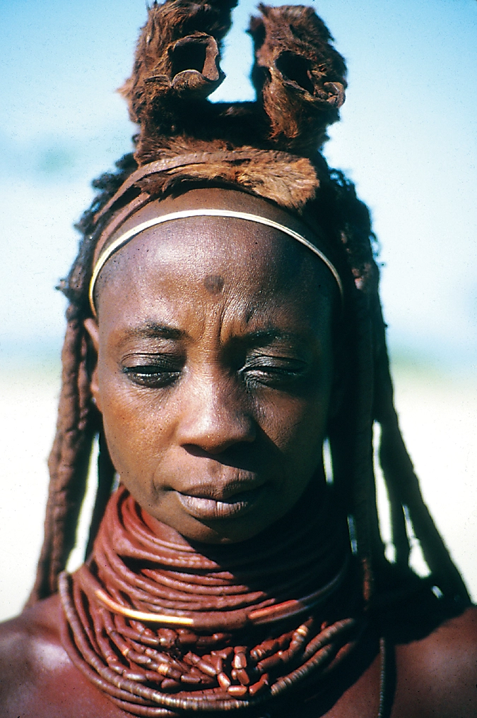 A Himba woman with a black dot in the middle of her forehead, an animal skin headdress, and multiple thing necklaces.