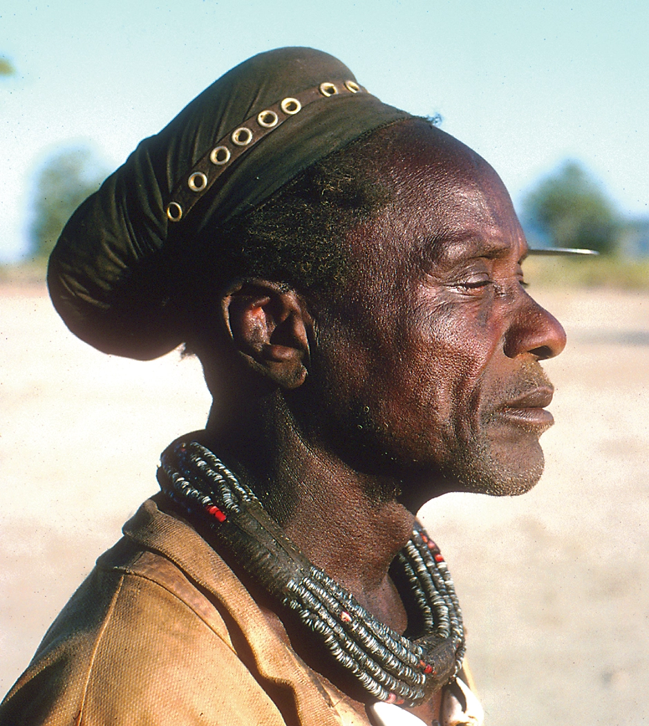A Himba man in a cap that covers his hair with a large, coiled necklace.