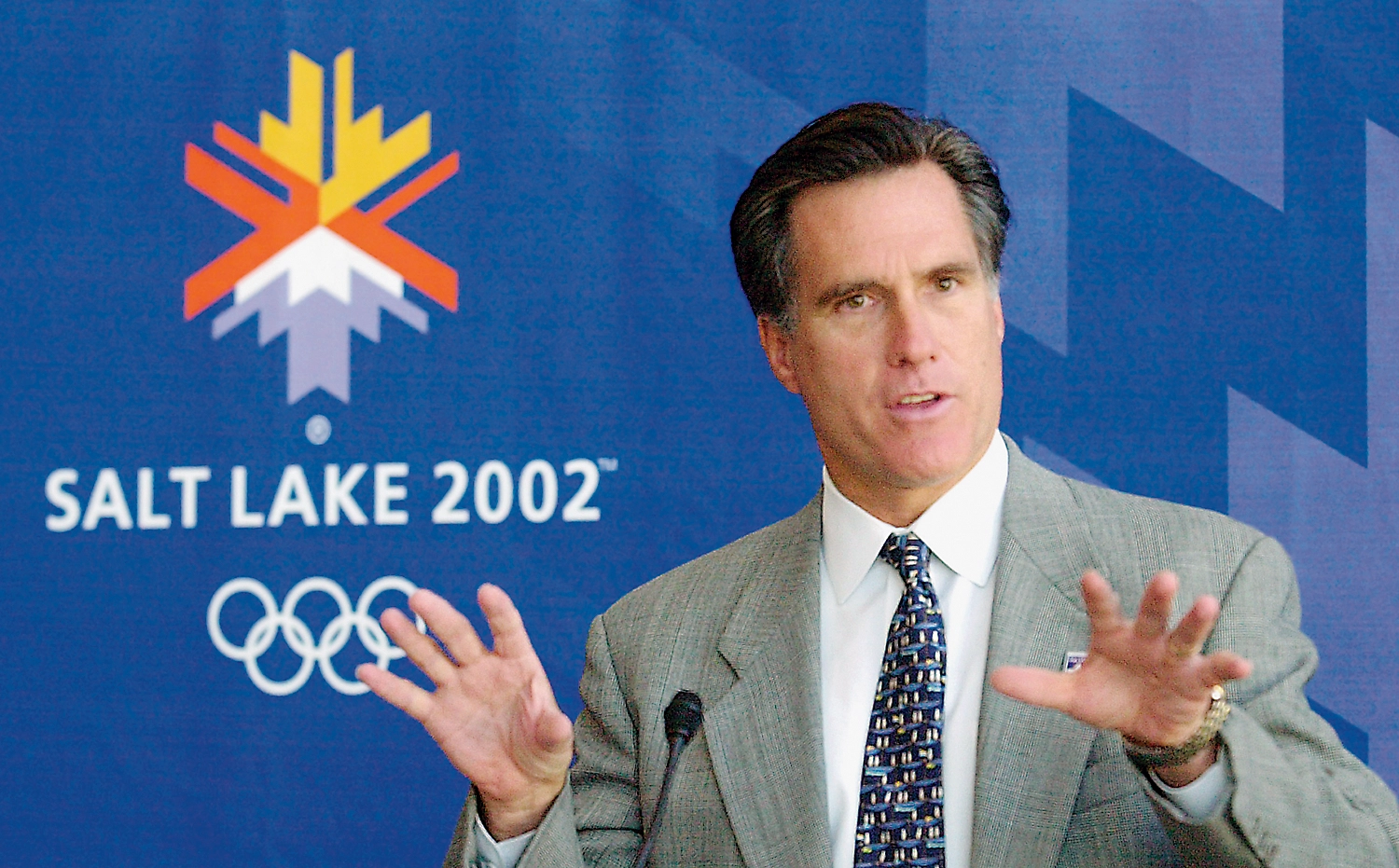 Mitt Romney speaking in front of a sign that reads "Salt Lake 2002" with the Olympic rings beneath.