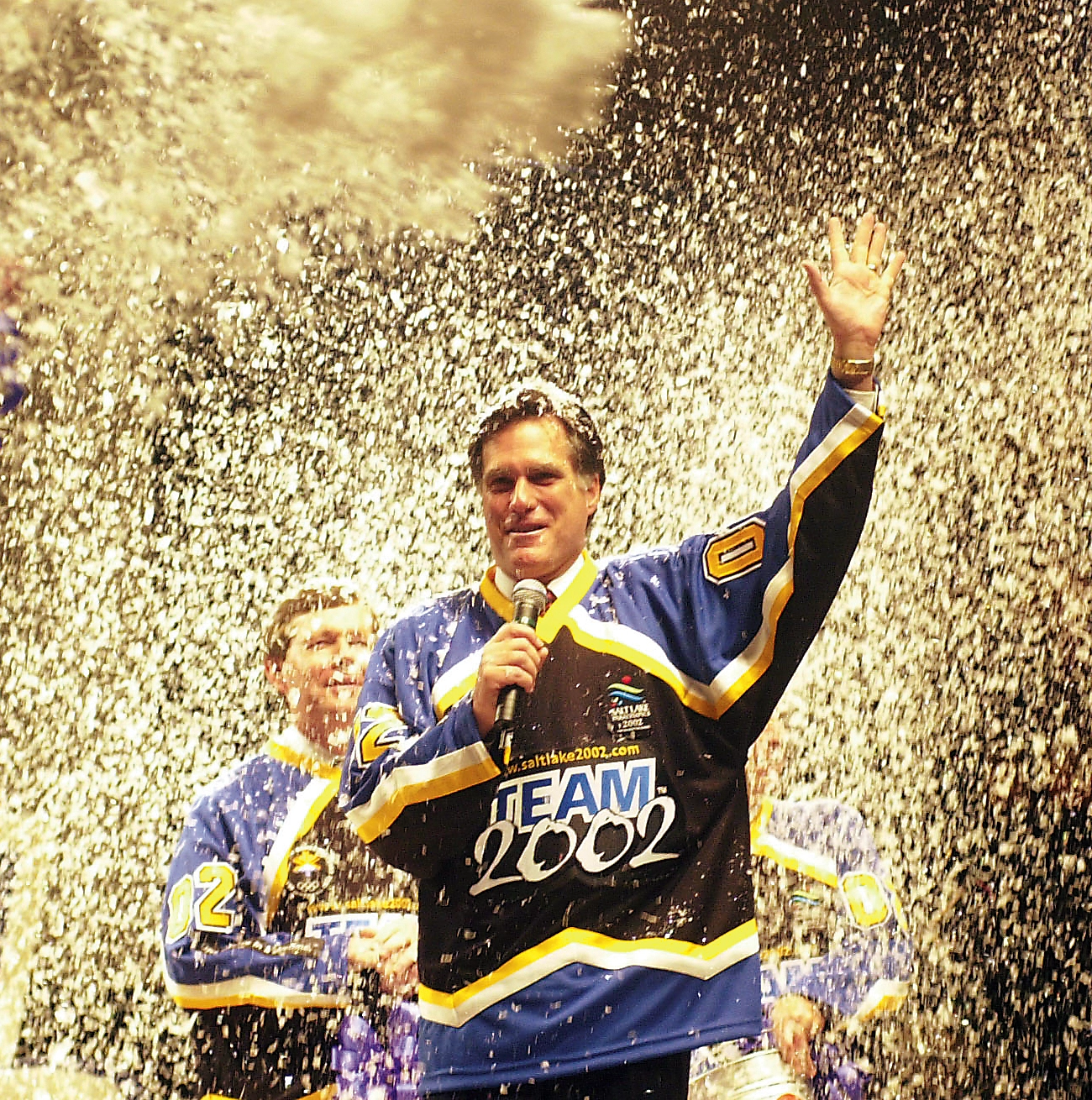 Mitt Romney stands holding a microphone while waving to a crowd while getting showered with snow.