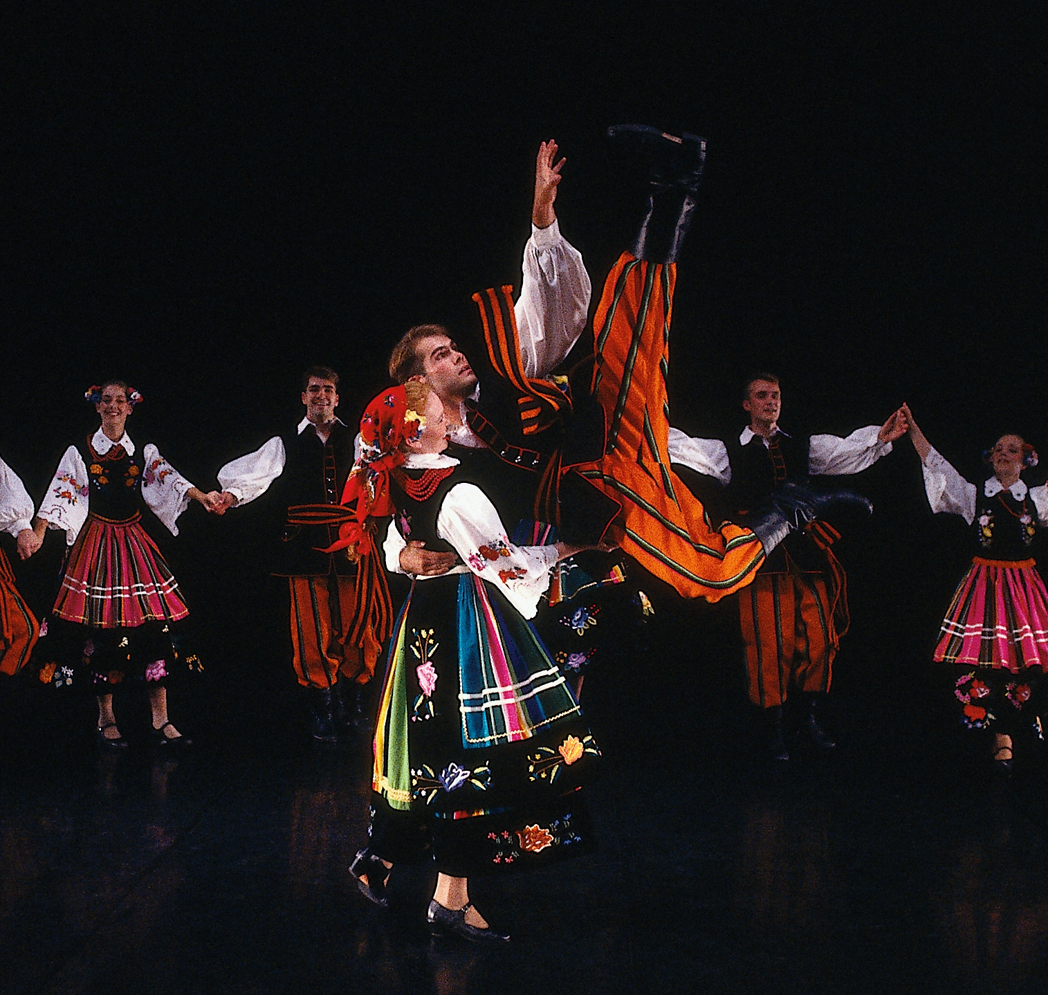 A man in traditional Polish dress performs a high kick whole holding on to his partner.