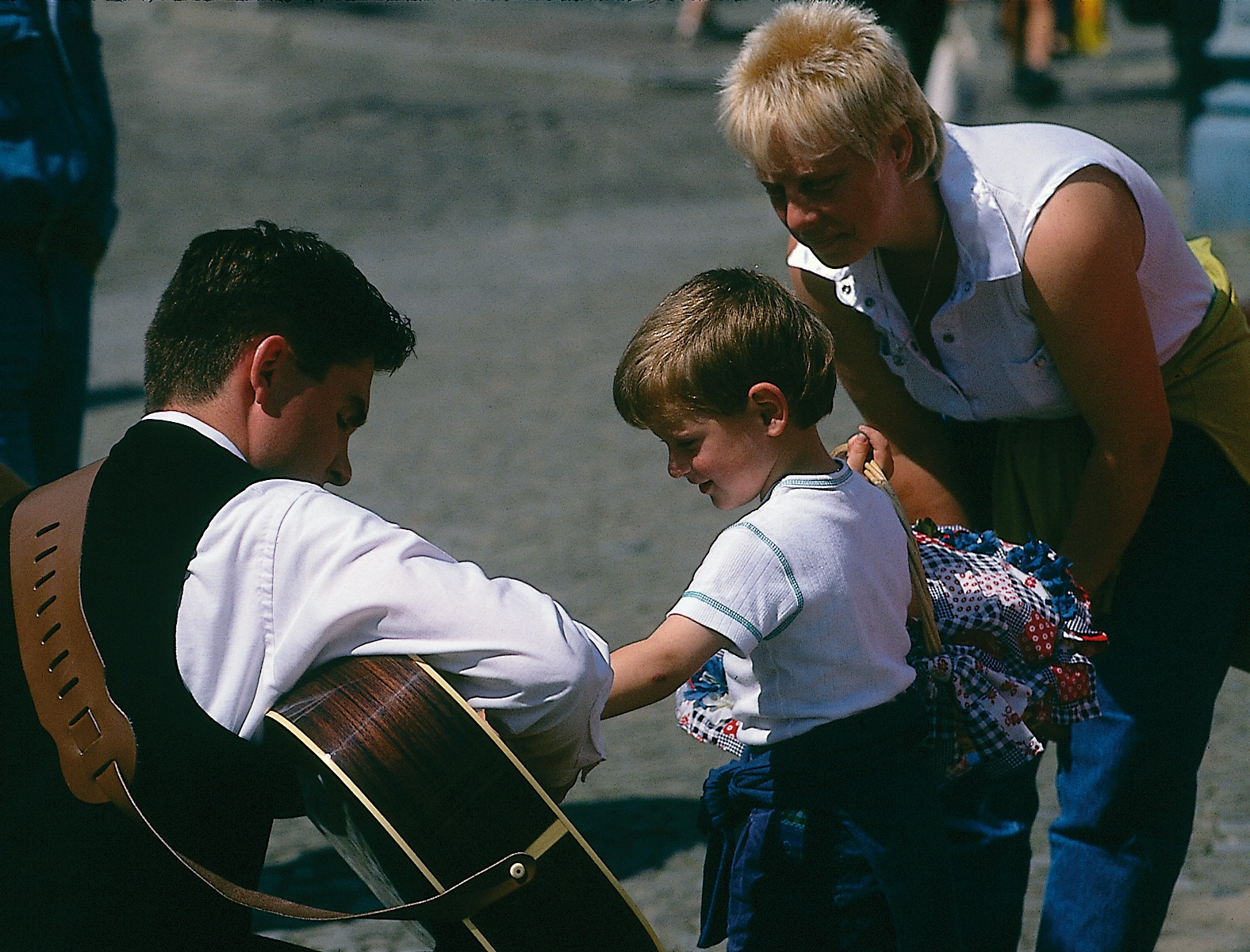 A man kneels down to let a child try his guitar while the boy's mother observes from behind.