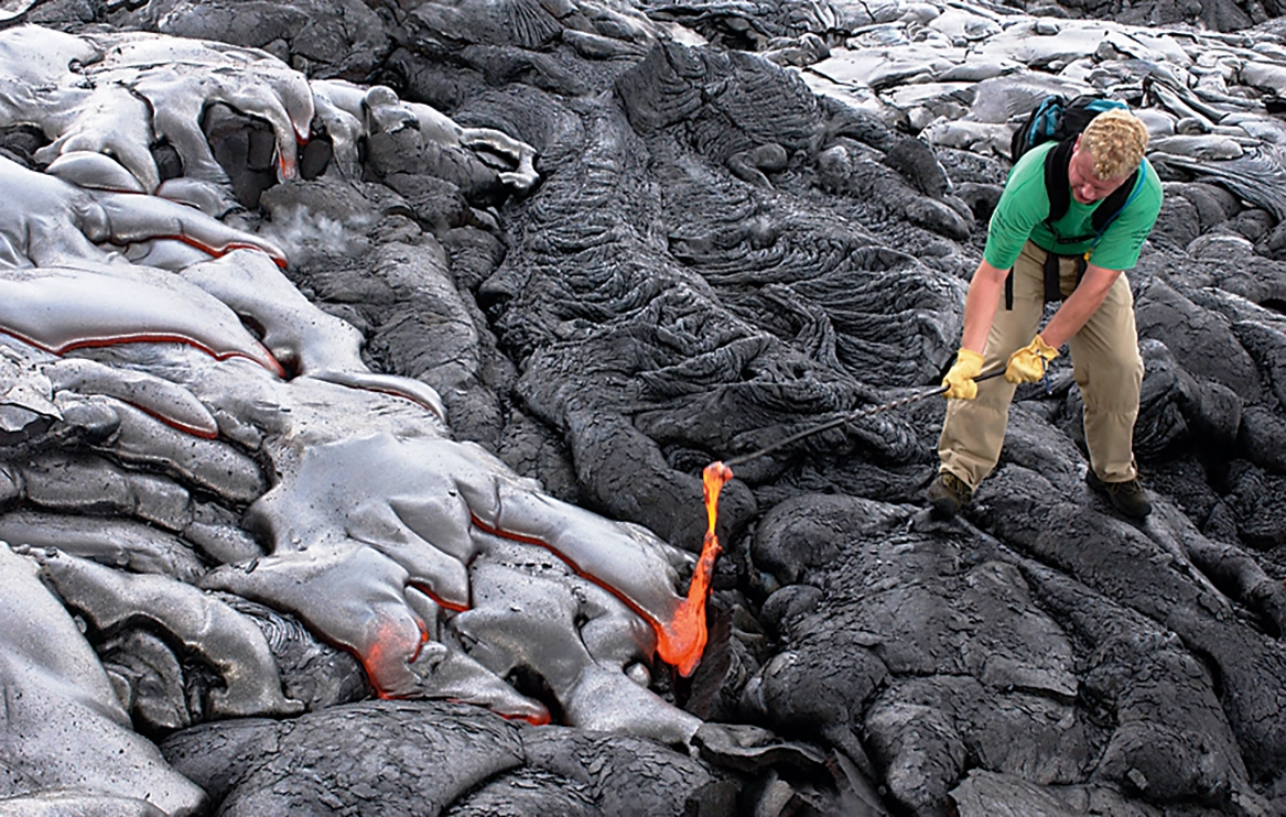 Matthew P Harper stands on a lava flow using a rod to pull a lava sample from a recent flow.