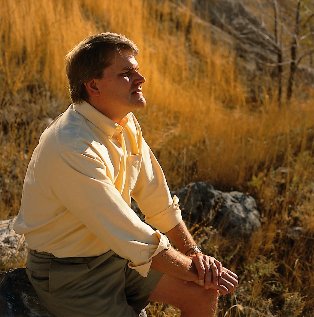David Crandall sits on a rock in a grassy field with his eyes closed in thought.