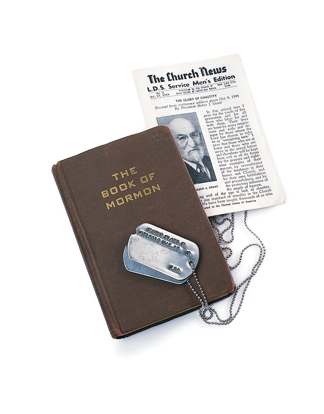 An older print edition of the Book of Mormon with a pair on dog tags on top and a news paper reading "The Church News: LDS Service Men's Edition" behind it.