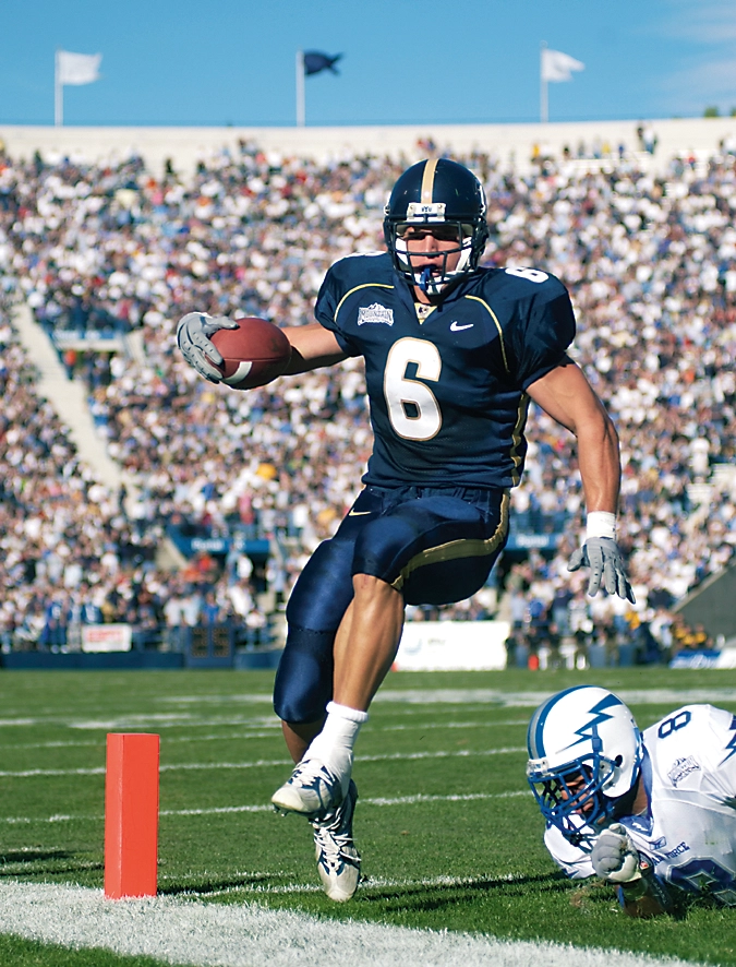 A football player in blue jumps into the air with the ball in hand to dodge an opposing player