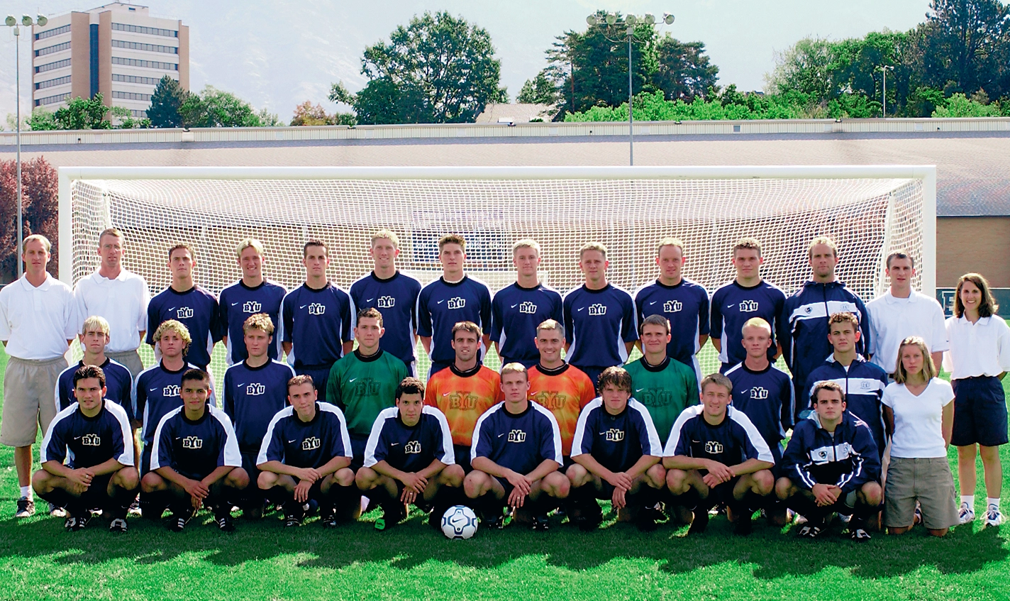 Two rows of soccer players pose with their coaches in front of a goal post.