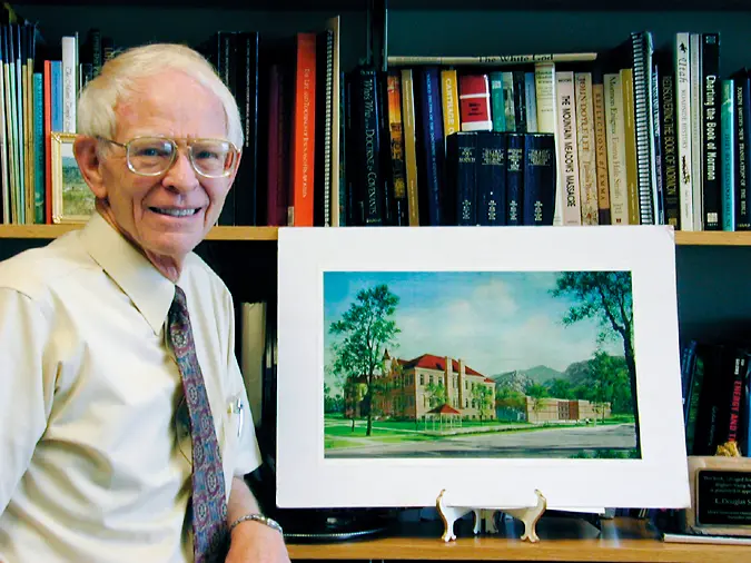 Douglas Smoot sits beside a bookshelf, smiling toward the camera, with a framed watercolor of the restored Brigham Young Academy building displayed in front of him.
