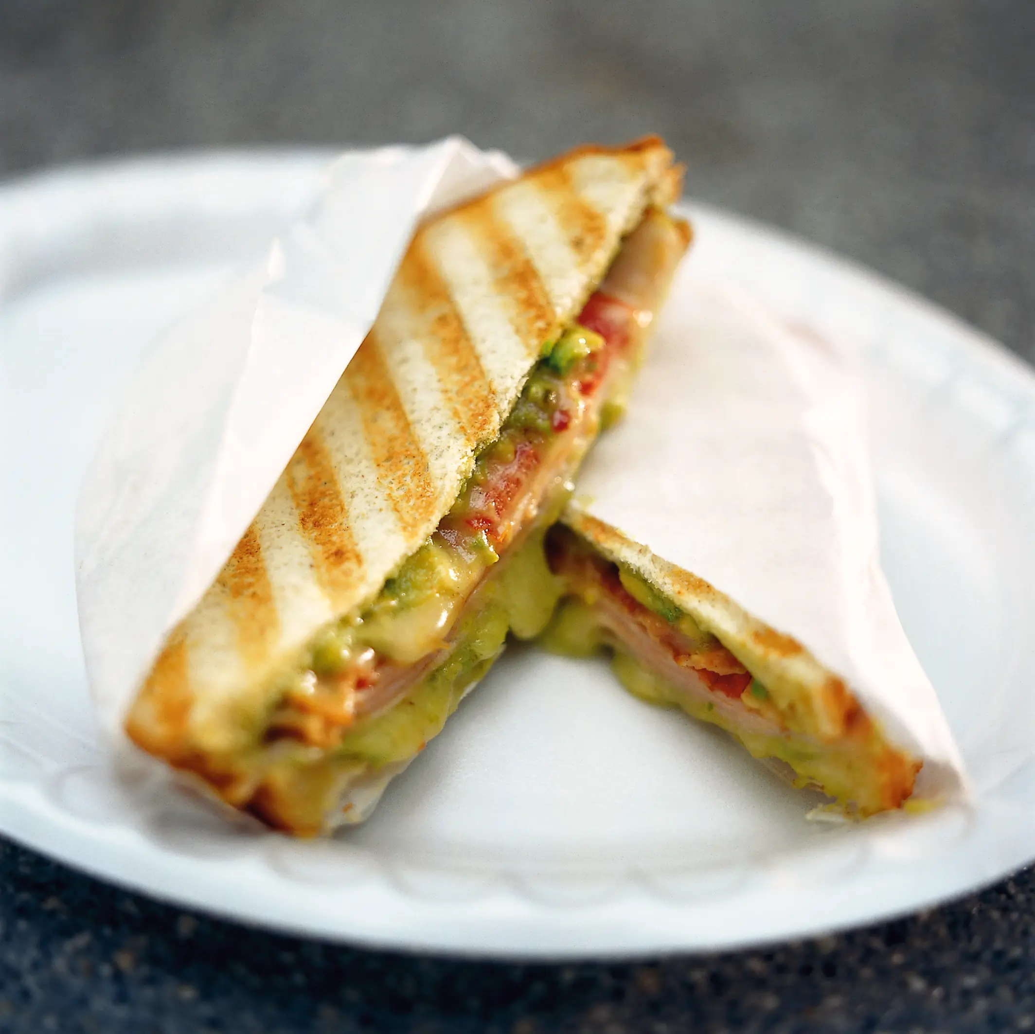 Grilled Montana panino cut into two triangular halves, with melted cheese, guacamole, tomato, turkey, and bacon visible between toasted bread.