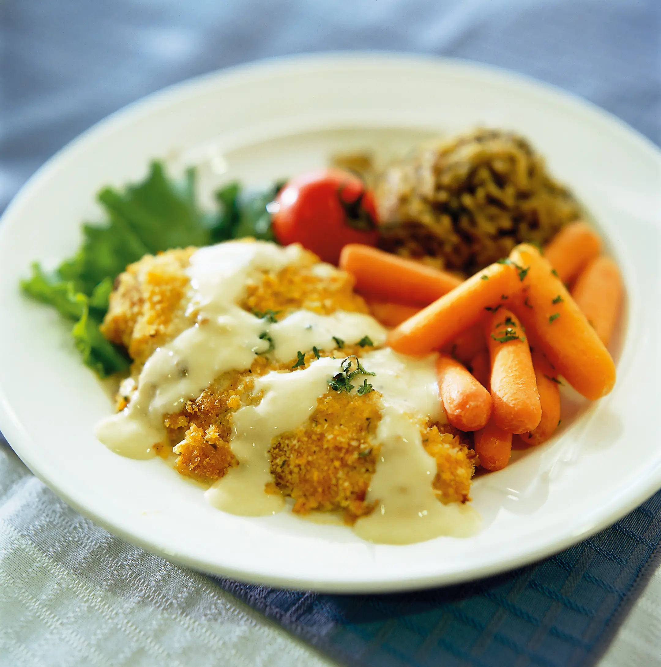 Herb-crusted chicken served with glazed baby carrots, leafy greens, a cherry tomato, and a mound of rice on a white plate.