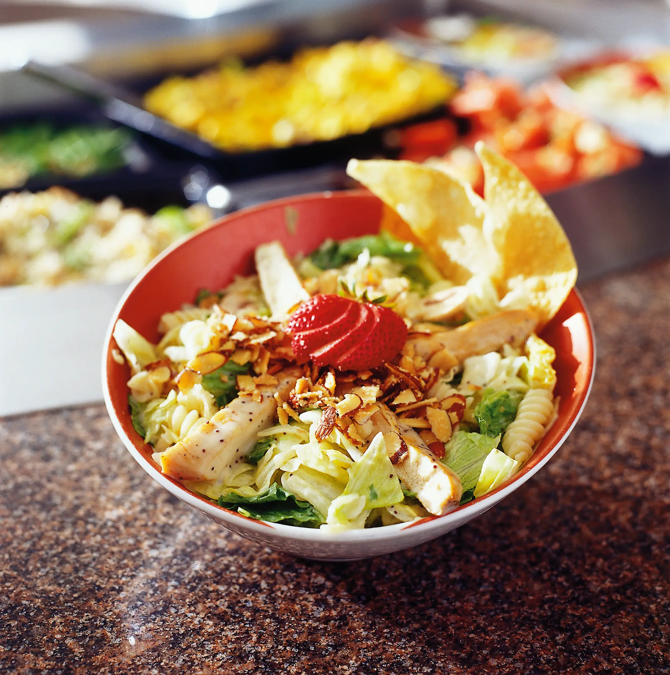 Bowl of pasta poppyseed salad with leafy greens, spiral pasta, sliced chicken, strawberries, almonds, and tortilla chips.