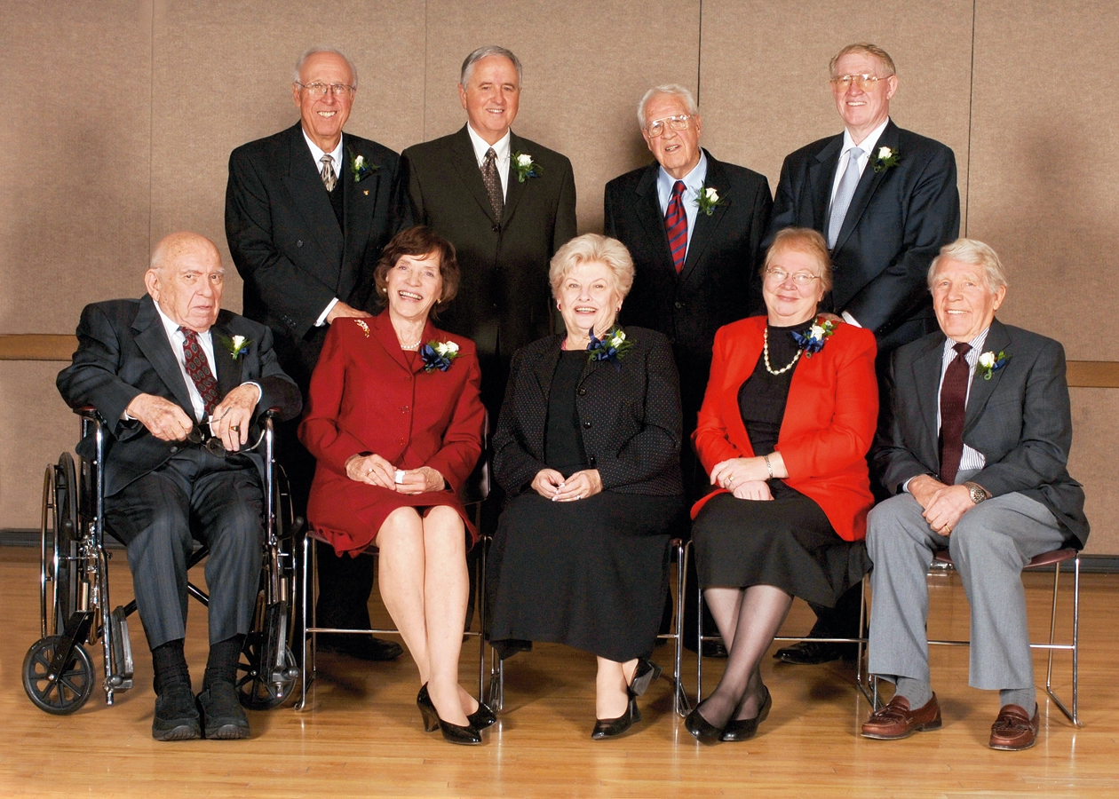 A row of four men in suits stands behind a row of two men and three women seated in front.