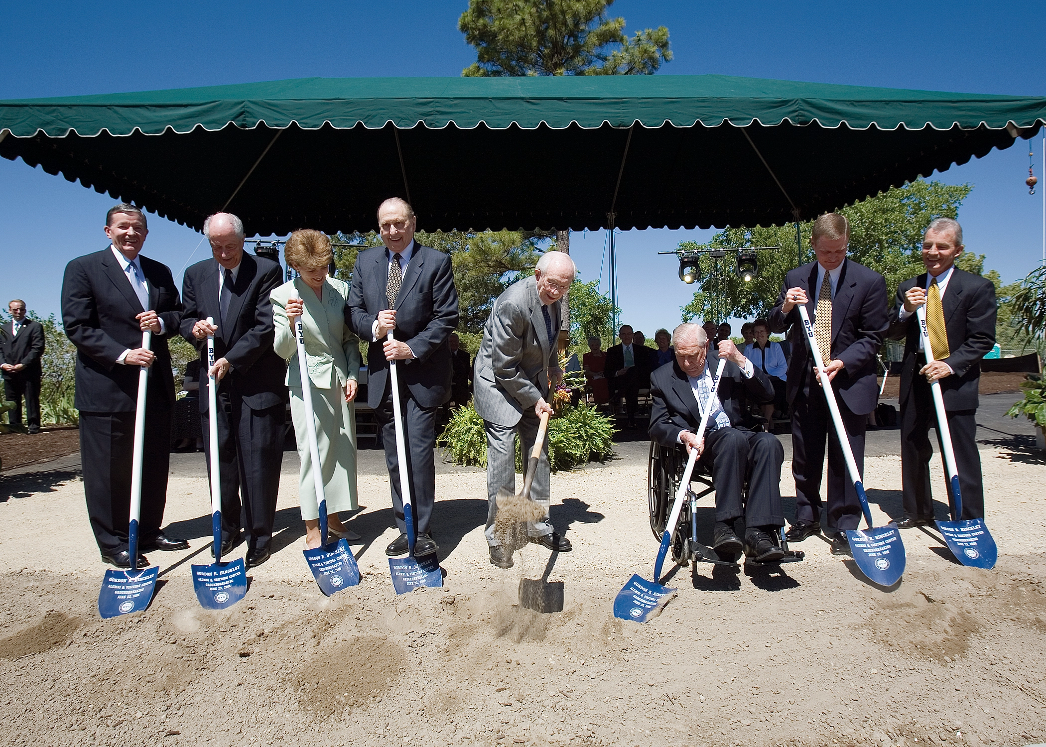A group of seven men and one women in suits place shovels into the dirt to begin the groundbreaking for the building.