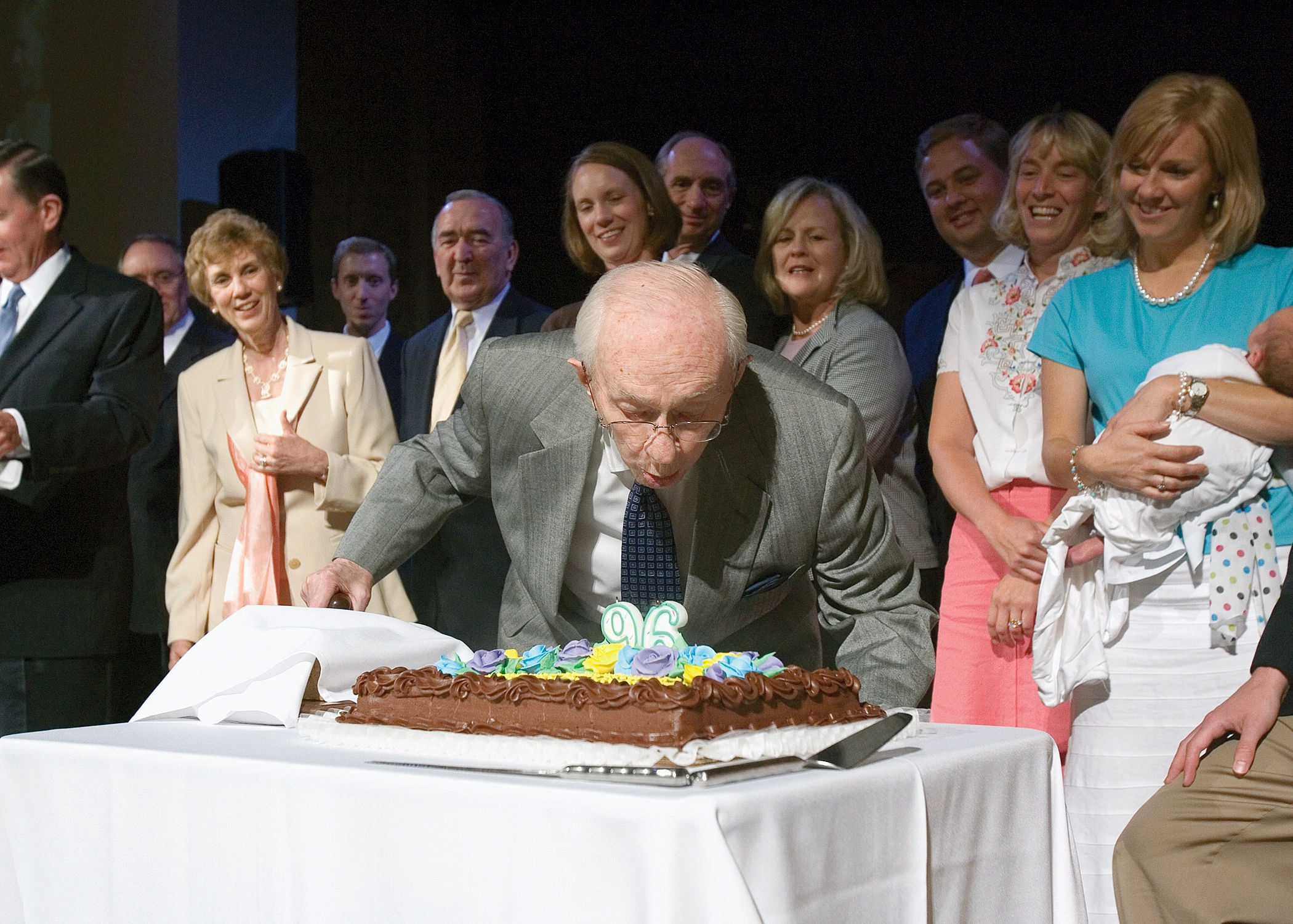 President Hinckley blows out candles in the shape of a 96 on a cake amidst a group of onlookers.