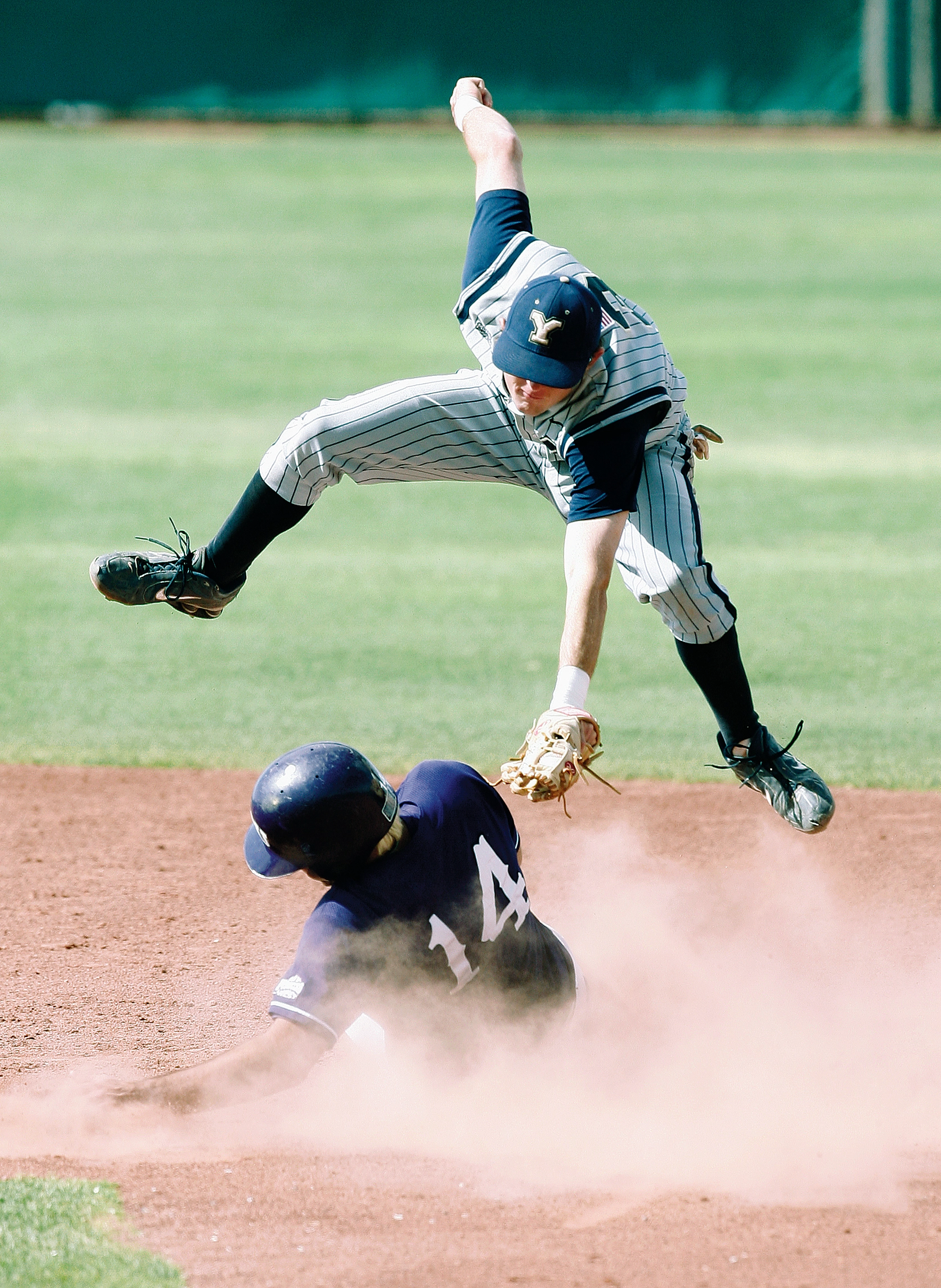 A baseball player leaps into the air to tag another player as he slides to the plate.
