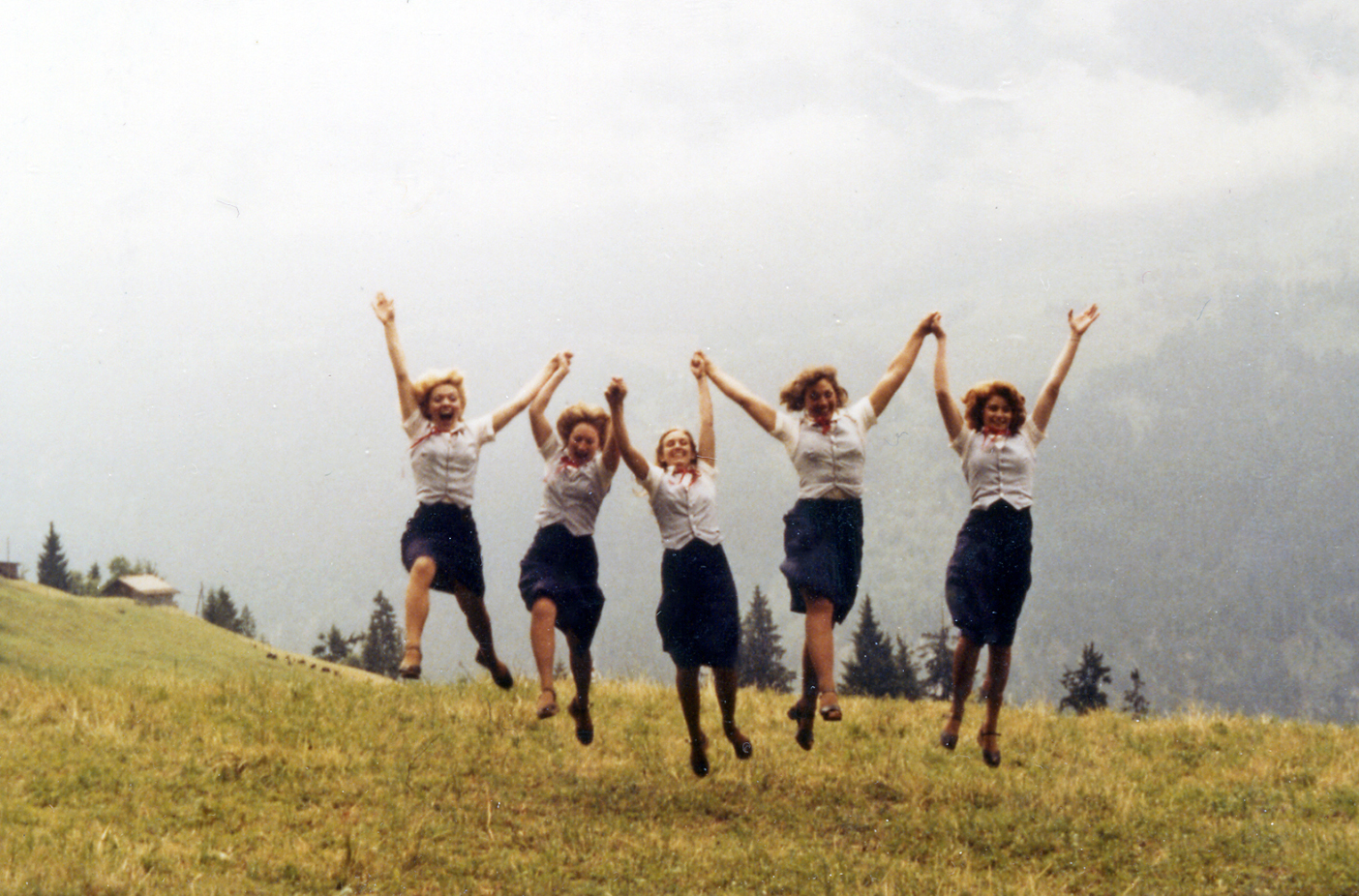 Five BYU folk dancers jump in the air with their arms up on a grassy hill.
