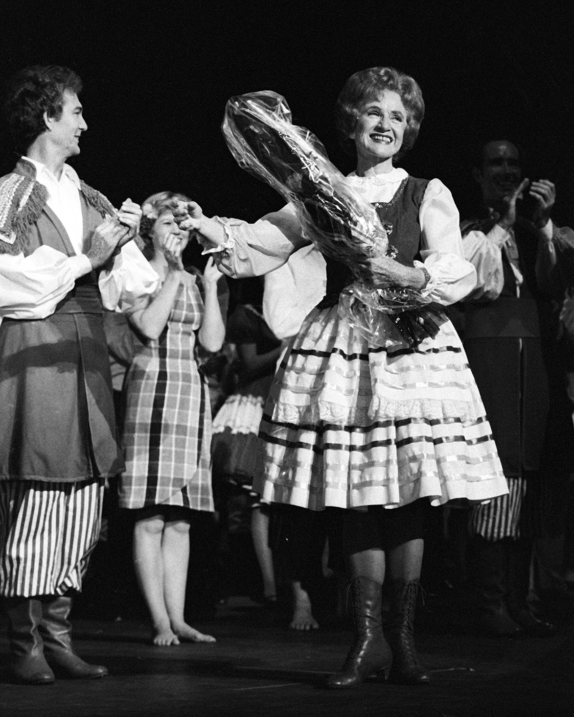 Mary Bee Jensen stands on stage holding a bouquet of flowers while performers stand around and clap.