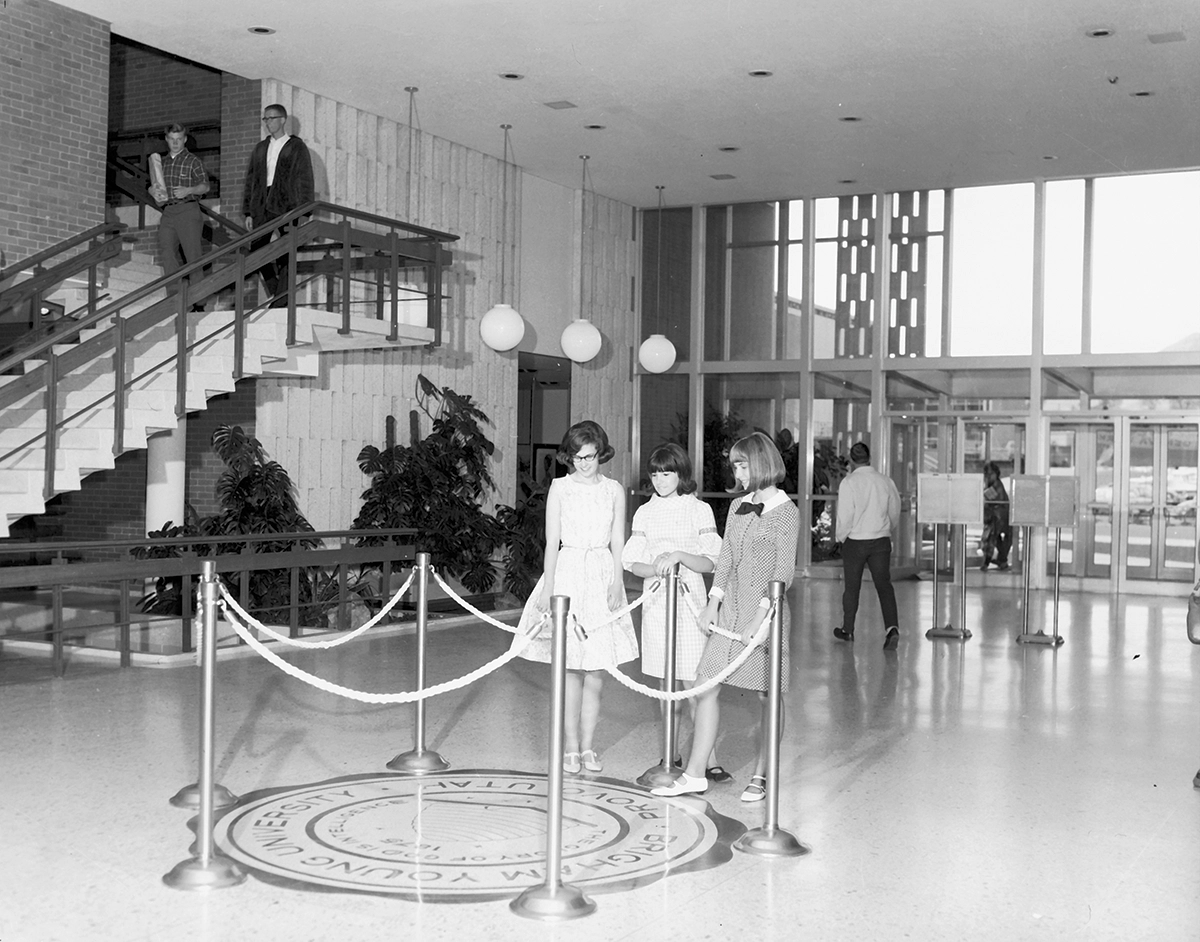 Three women stand in front of a roped off floor section bearing the seal of BYU
