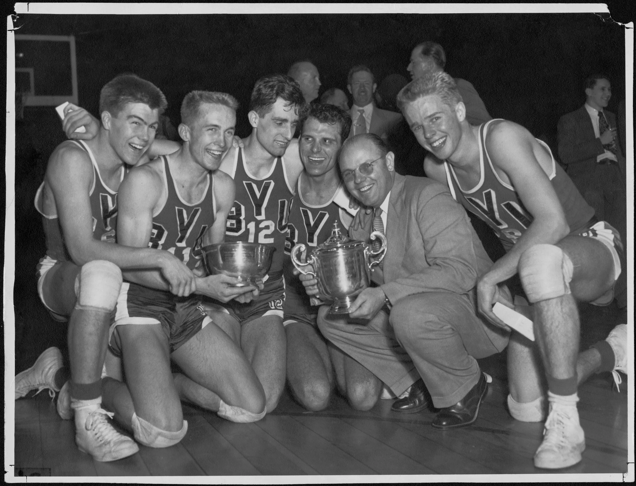 Six basketball players in BYU uniforms kneel with their coach while holding a trophy.