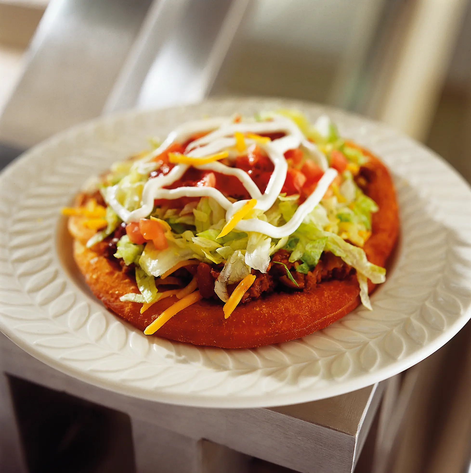 Close-up of a Navajo taco with thick fry bread topped with beef chili, shredded cheddar cheese, lettuce, diced tomatoes, salsa, and ranch dressing.