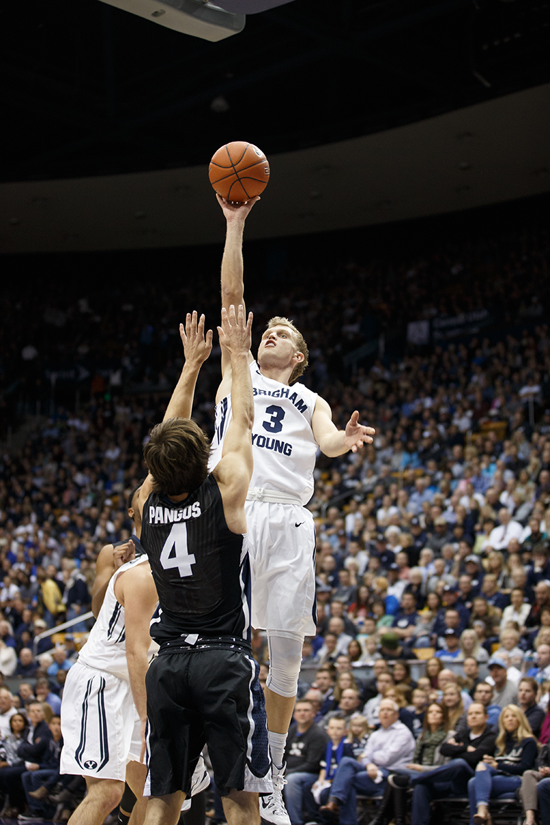 Tyler Haws jumping, about to release the ball while a member of the other team reaches up to block him.
