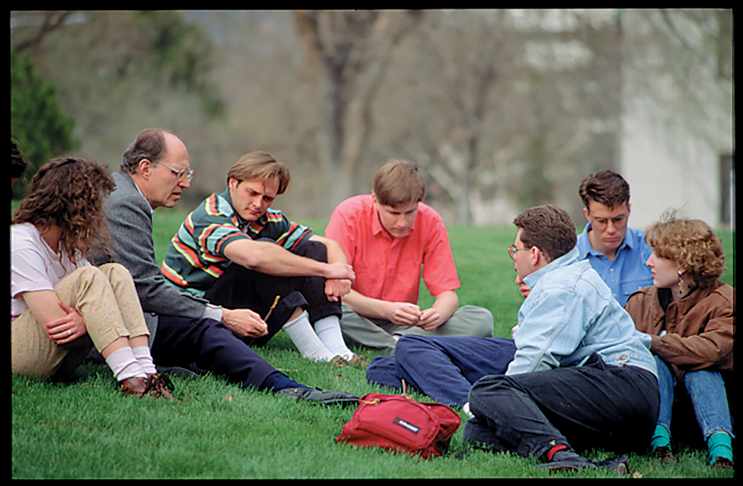 A few students gathered around on the grass surrounding their teacher as he talks to them.