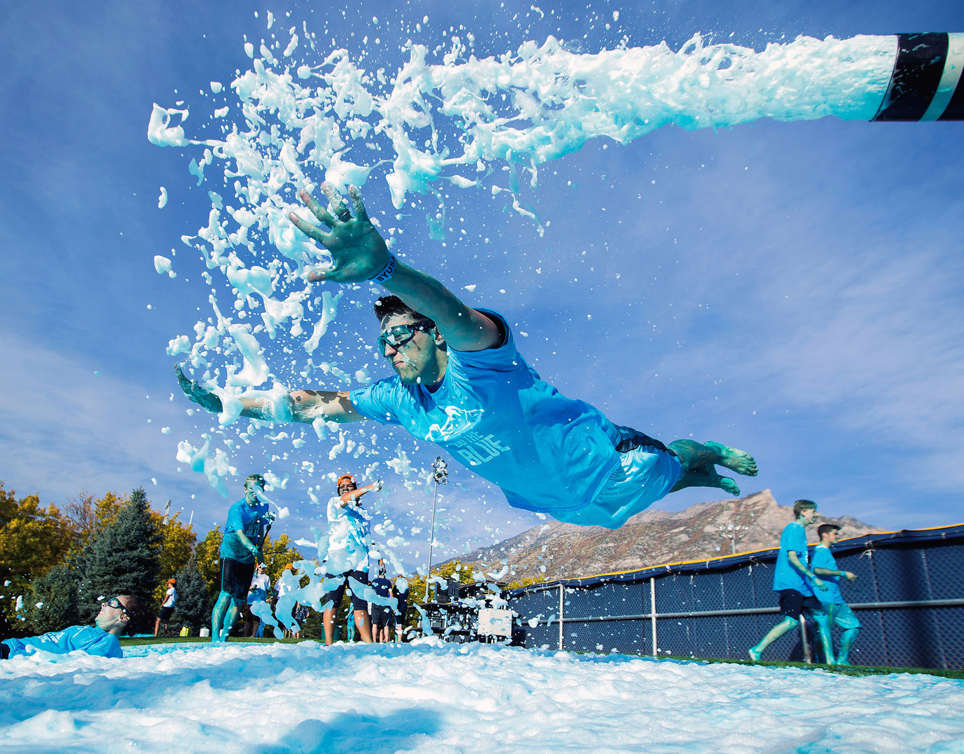 A student dives into a slide of blue foam as a hose sprays more on him