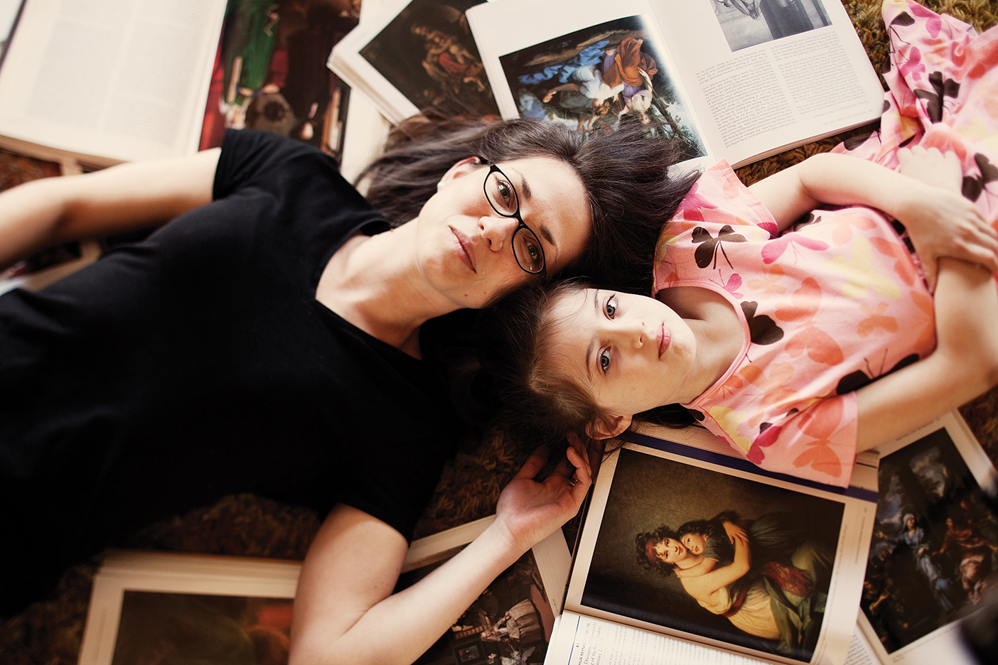 mother and daughter smile at the camera while laying down on the ground surrounded by pictures and books