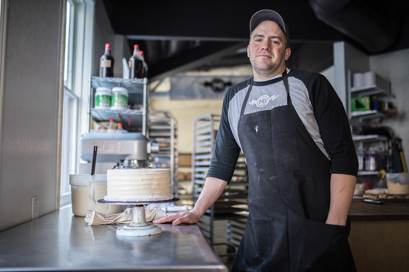 A photograph of Peter Tidwell in his bakery standing next to a completed, frosted cake.