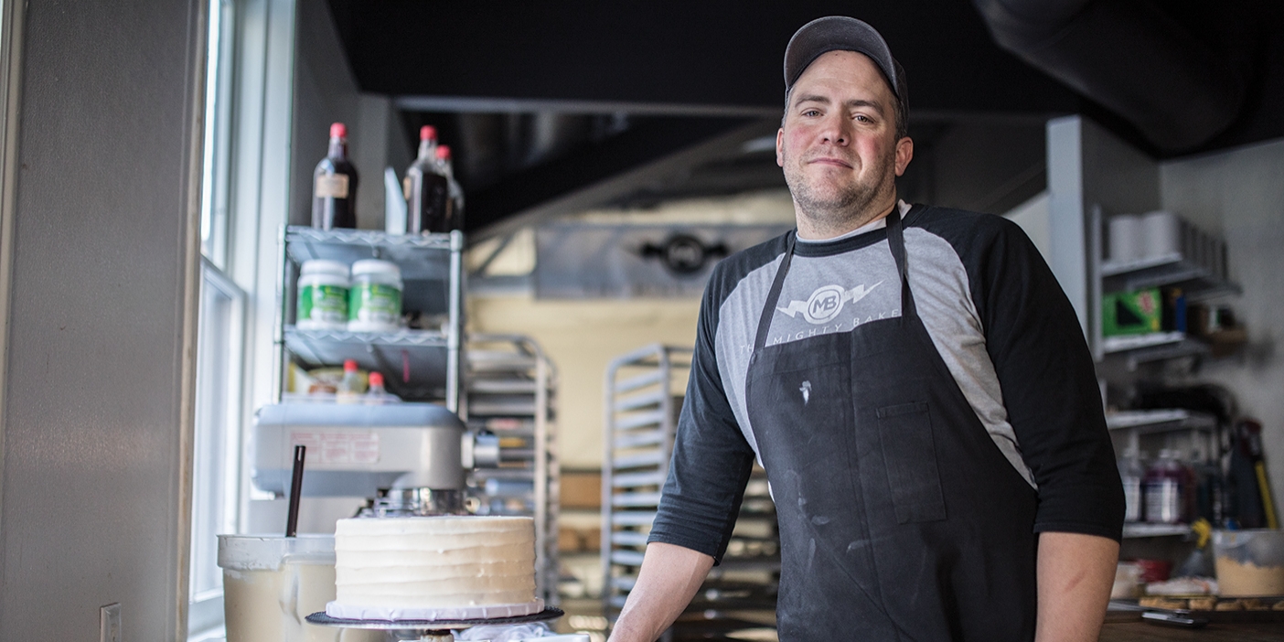 A photograph of Peter Tidwell in his bakery standing next to a completed, frosted cake.