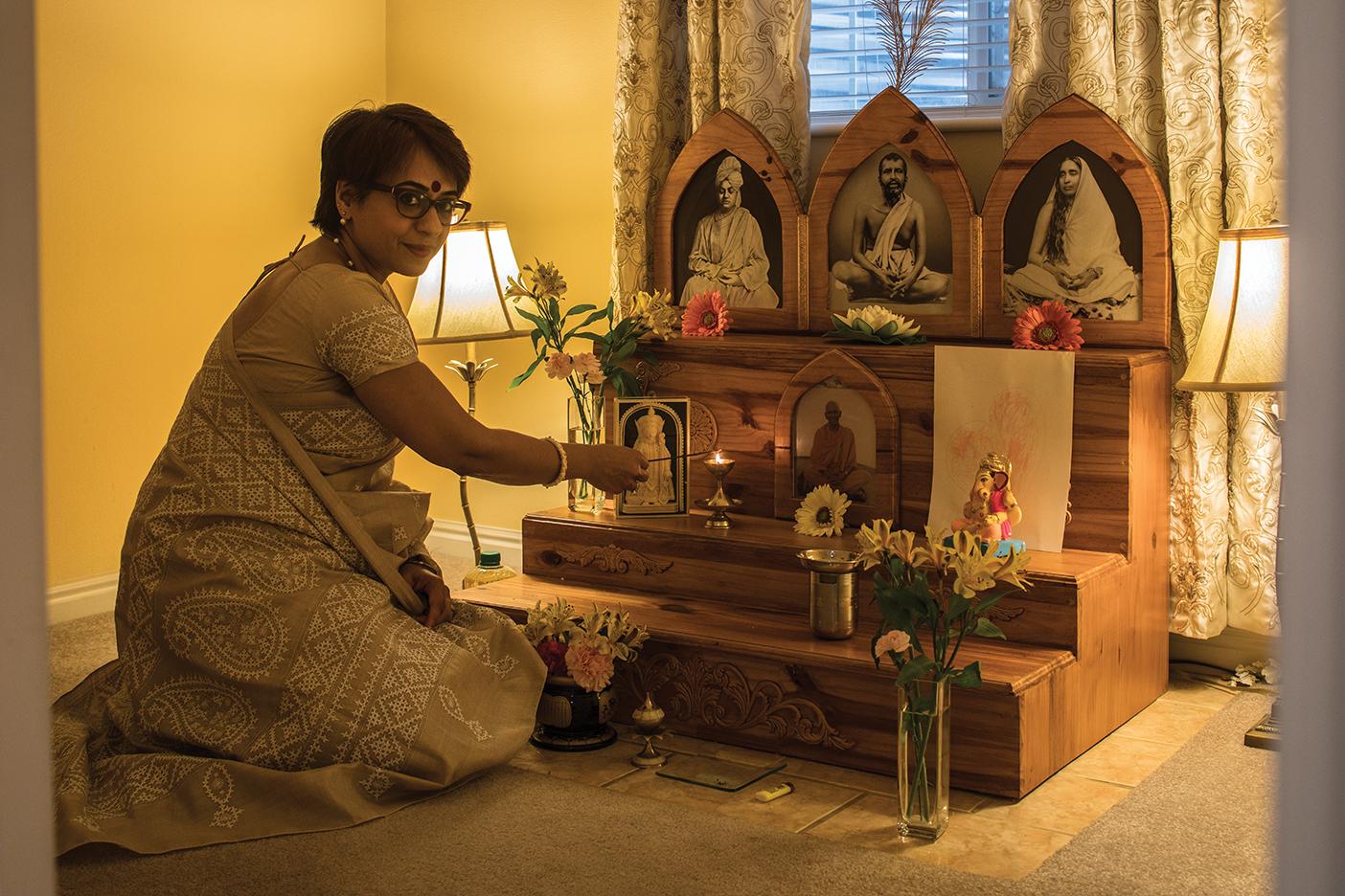 Pritha Lal, dressed in traditional Hindu clothing kneels in front of a family shrine in her home that displays pictures of family members
