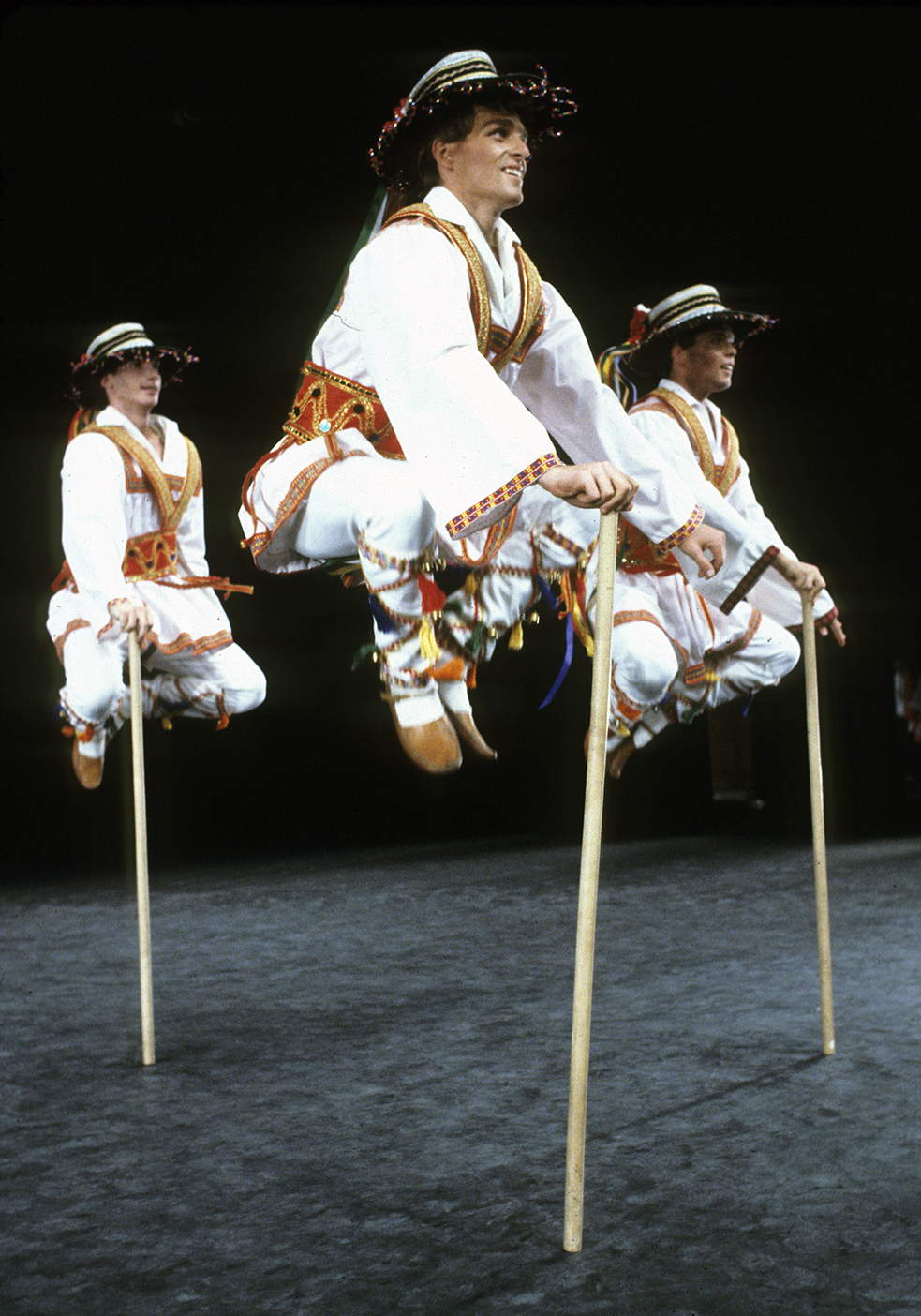 Three male folk dancers wearing traditional costumes jump high into the air during a dance on stage