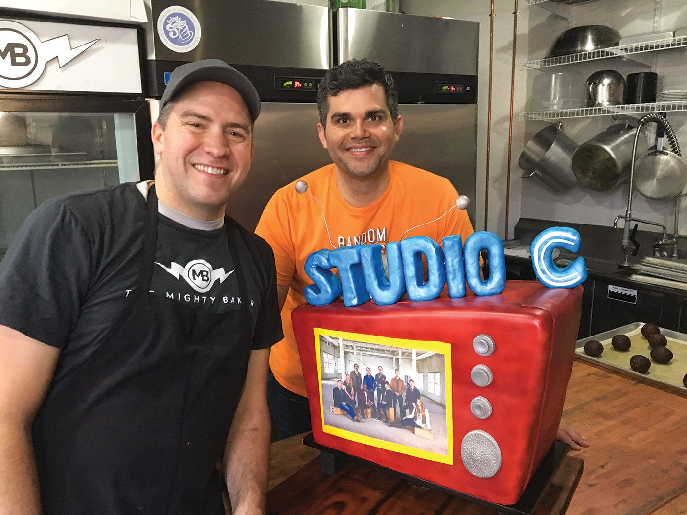 Peter Tidwell and host of TV show Random Acts pose with a large red Studio C-themed cake that is shaped like a TV