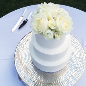 a photo of a white, three-tiered cake that is topped with white roses and displayed on a table with a white tablecloth
