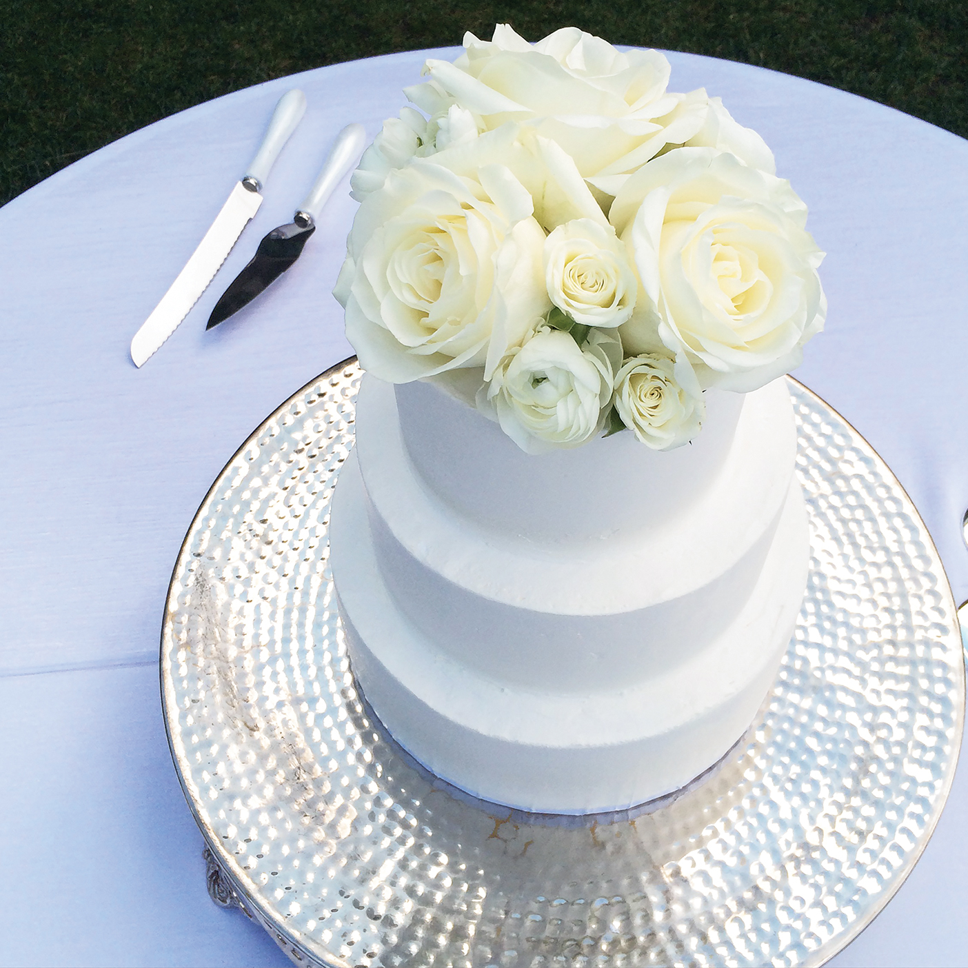 a photo of a white, three-tiered cake that is topped with white roses and displayed on a table with a white tablecloth