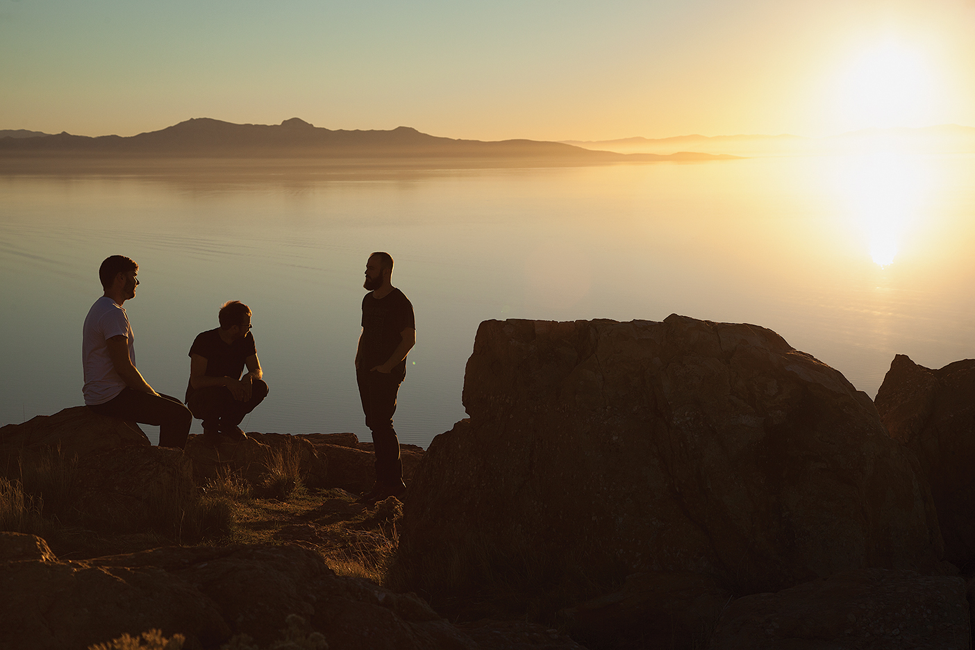 band members sit on rocks near water as the sun sets