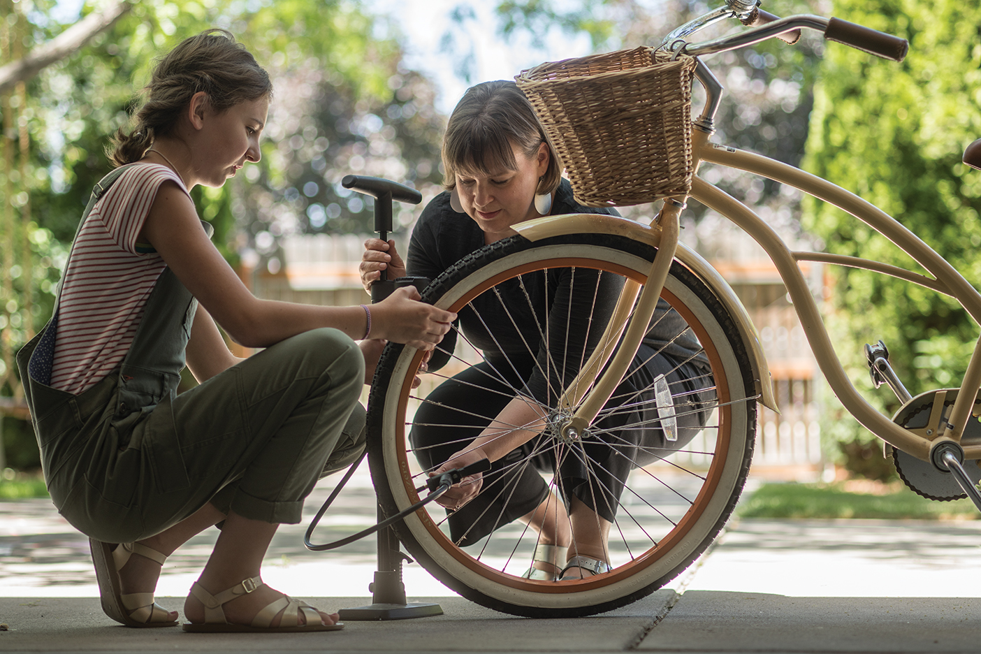 A mother bends down as she helps her daughter pump up a bicycle tire.