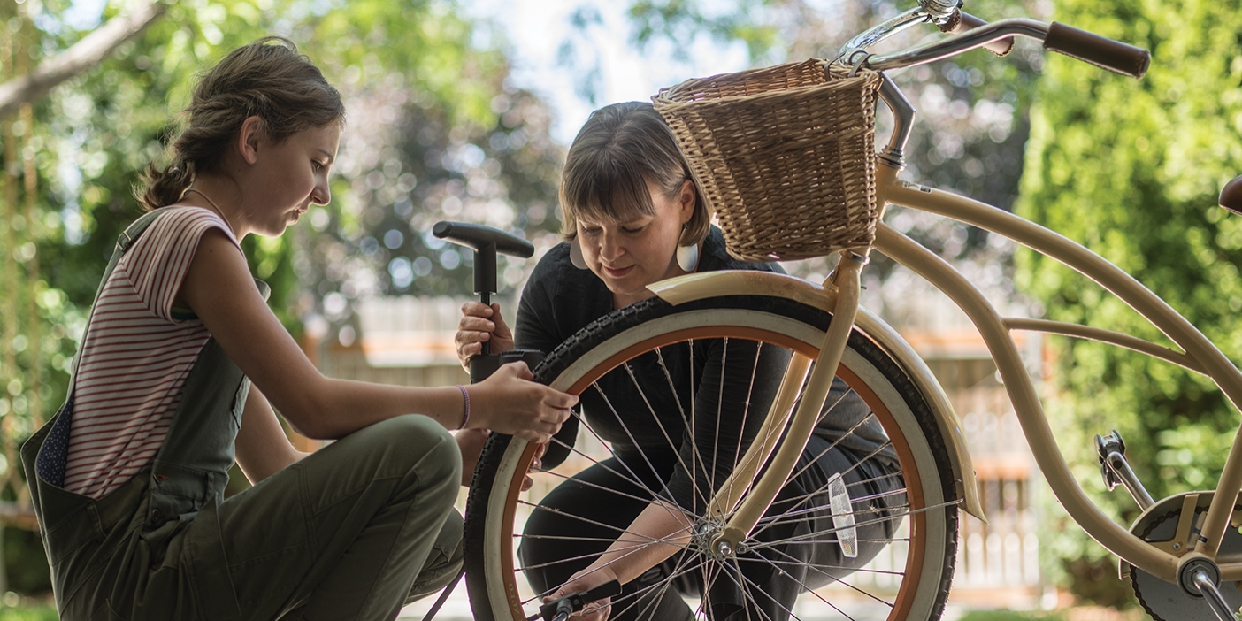 A mother bends down as she helps her daughter pump up a bicycle tire.