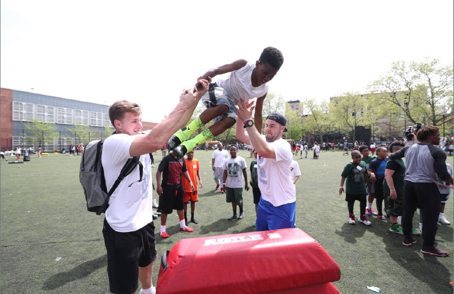 A participant leaps over an obstacle with the help of two BYU players.