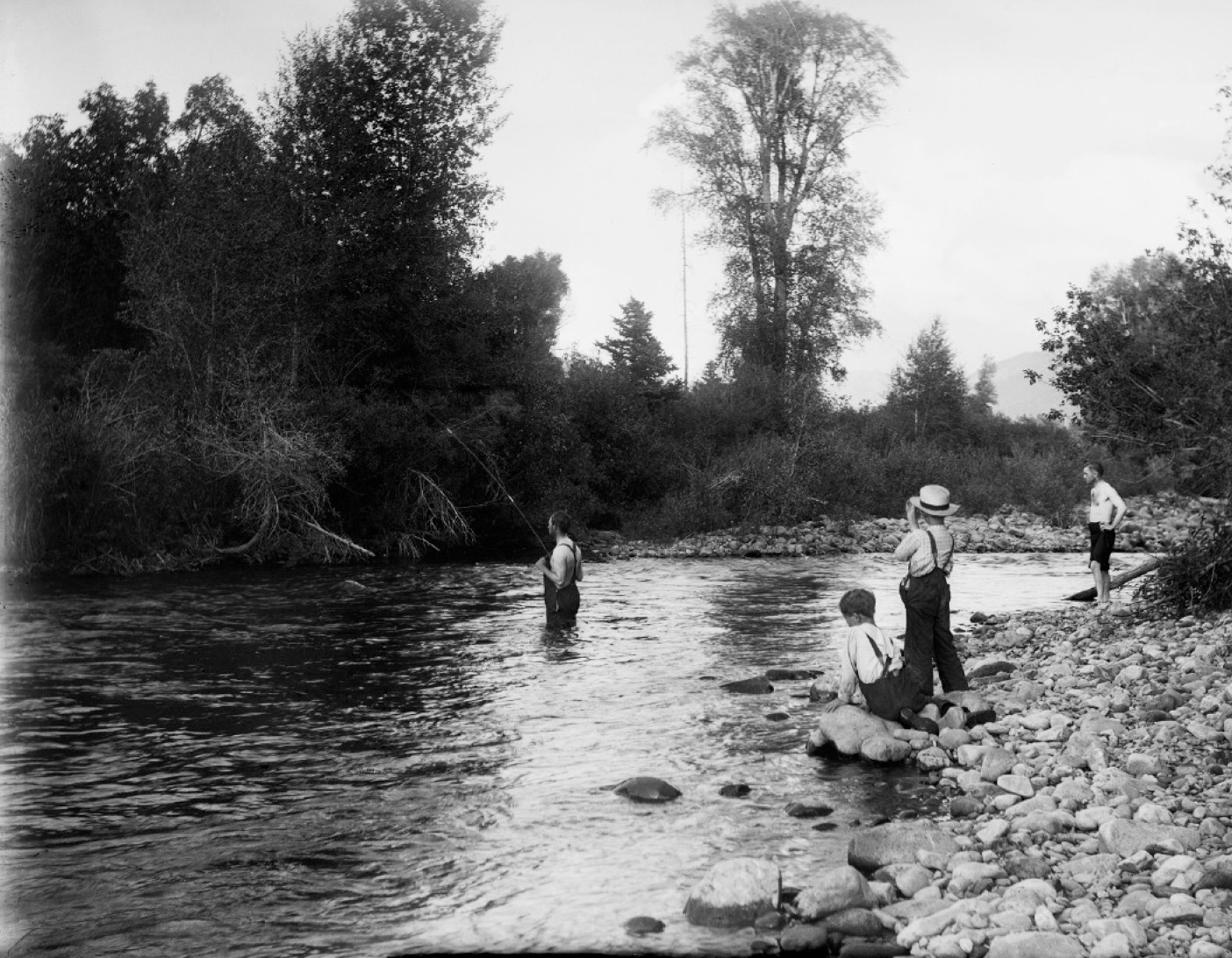 A black and white photo of young boys fishing and wading in a river